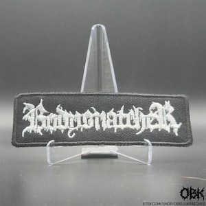 May include: Black rectangular patch with white embroidered text. The text appears to be a band name or logo. The patch is displayed on a clear plastic stand against a dark background. The Etsy shop name is visible in the bottom right corner.