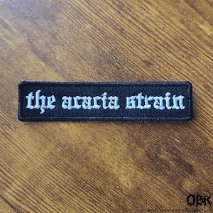 Può includere: Una toppa rettangolare nera ricamata con il testo "the acacia strain" in un carattere bianco in stile gotico. La toppa presenta un bordo cucito ed è progettata per essere applicata su articoli in tessuto, con lettere bianche su sfondo scuro.