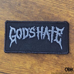 May include: A rectangular black patch with the word "GODSHATE" embroidered in gray, stylized, gothic lettering. The patch has a black border and is set against a wooden surface. The Etsy shop URL is visible in the bottom right corner.