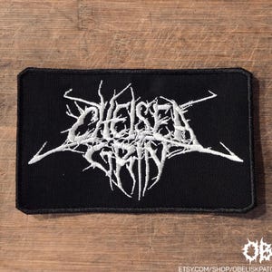 Puede incluir: Un parche rectangular bordado en negro que presenta el texto blanco Chelsea Grin en una fuente estilizada, dentada y similar a ramas. El parche tiene un borde negro grueso y una superficie de tela texturizada con el texto centrado.