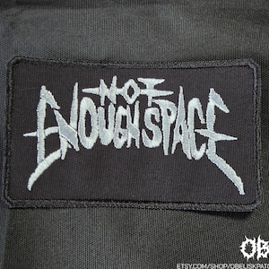 May include: A rectangular black embroidered patch featuring the text NOT ENOUGH SPACE in a stylized, jagged, light gray font. The patch has a black stitched border and is set against a black fabric background with a small logo in the corner.