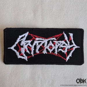 May include: A rectangular black embroidered patch featuring white stylized lettering that reads Cryptopsy. The text is layered over a thin, red, web-like or star-shaped embroidered design. The patch has a black stitched border and a flat finish.