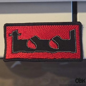 May include: Embroidered patch featuring the word "TOOL" in a stylized black font, outlined in white, set against a red background. The patch has a black border. The patch is rectangular.
