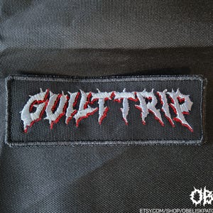 Op de afbeelding: Een rechthoekige zwarte geborduurde patch met de tekst GUILT TRIP in een gestileerd, grillig, zilvergrijs lettertype met scherpe rode accenten. De patch heeft een dikke zwarte gestikte rand en ligt op een achtergrond van zwarte textuurstof.
