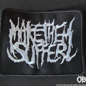 Puede incluir: Un parche rectangular negro con texto bordado en blanco que dice "MAKE THEM SUFFER" en una fuente gótica. El parche es para ropa o accesorios.