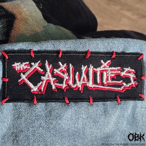Puede incluir: Un parche rectangular negro con el texto "THE CASUALTIES" en blanco y rojo, cosido sobre tela vaquera azul claro. El parche está bordeado con costuras rojas. La imagen es de un parche de punk rock.