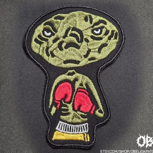 Puede incluir: Un parche bordado que presenta una cabeza de criatura verde estilizada con una superficie texturizada sobre un torso que lleva guantes de boxeo rojos, una cinturilla blanca y pantalones cortos amarillos, todo delineado en hilo negro.