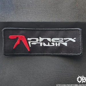 Può includere: Una toppa rettangolare nera ricamata con un logo rosso Aphex Twin sul lato sinistro e una scritta bianca ricamata che recita Aphex Twin sul lato destro. La toppa presenta un bordo cucito ed è posizionata su un tessuto scuro testurizzato.