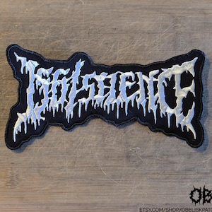 May include: A black embroidered patch featuring white text that reads "ISOLENCE" in a stylised, jagged, dripping font. The patch has a distinct, irregular, and contoured border that follows the shape of the lettering, creating a sharp, high-contrast design.