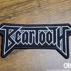 May include: A black rectangular patch featuring the word "Beartooth" in white, gothic lettering. The patch has a black border and is set against a wooden surface. The Etsy shop name is visible at the bottom.
