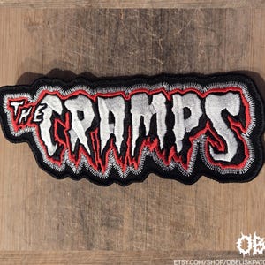 May include: Embroidered patch featuring the band name "THE CRAMPS" in white, outlined in red, with a black border. The patch has a jagged, dripping design, and is set against a wooden background. The patch is a music-themed accessory.