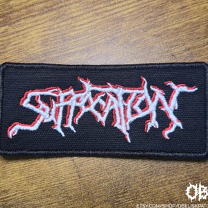 Può includere: Toppa rettangolare nera con la scritta "SUFFOCATION" ricamata in bianco con un contorno rosso. La toppa ha un bordo nero ed è probabilmente per abbigliamento o accessori. Il nome del negozio Etsy "OBK" è nell'angolo in basso a destra.