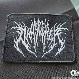 May include: Black rectangular patch with white embroidered text in a stylized, gothic font. The patch has a black border and is set against a dark background. The text is illegible.