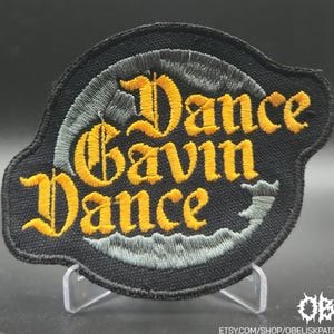 Könnte beinhalten: Schwarzer, gestickter Aufnäher mit den Worten "Dance Gavin Dance" in gelben, stilisierten Buchstaben. Der Aufnäher hat einen schwarzen Rand und ein grau gesticktes Design. Der Aufnäher wird auf einem transparenten Ständer präsentiert.