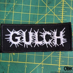 May include: A black rectangular patch with the white embroidered word "GULCH". The letters have a spiky, stylized design. The patch is on a green surface with grid lines.