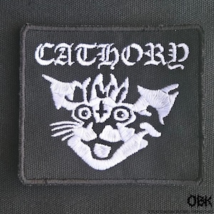 May include: Black square patch with white embroidered text "CATHORD" above a stylized cat face. The cat face has large eyes, a wide grin, and pointed ears. The patch is likely for clothing or accessories.