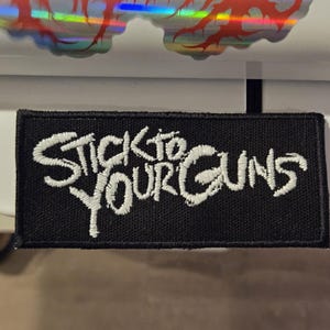 May include: Black rectangular patch with the words "STICK TO YOUR GUNS" embroidered in white, stylized lettering. The patch is likely for clothing or accessories.
