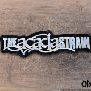 Puede incluir: Un parche bordado negro que presenta el texto THE ACACIA STRAIN en letras blancas estilizadas. El parche tiene un borde irregular y dentado que sigue el contorno del texto, con adornos decorativos blancos que se extienden desde las letras.