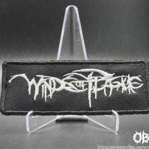 May include: A rectangular black embroidered patch featuring white stylized text that reads Winds of Plague. The patch has a thick black stitched border and is displayed on a clear plastic stand against a dark background.