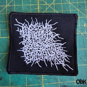 Puede incluir: Parche cuadrado negro con diseño abstracto bordado en blanco. El diseño es un grupo denso y enmarañado de líneas. El parche tiene un borde negro. La esquina inferior derecha tiene el texto "OBK" y una dirección web.