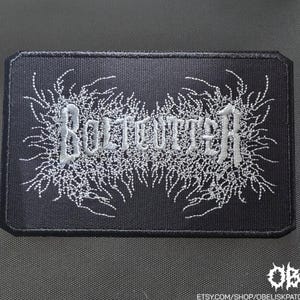 May include: Black rectangular patch with the word "BOLTOUTTER" embroidered in silver thread. The text is surrounded by a detailed, stylized, white floral design. The patch has a black border.