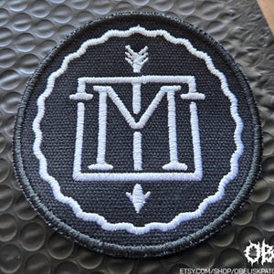 Könnte beinhalten: Runder, schwarzer Aufnäher mit weißem Stickdesign. Das Design enthält den Buchstaben "M" und dekorative Elemente, umrahmt von einem weißen, gewellten Rand. Wahrscheinlich ein Emblem oder Logo.