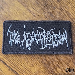 Puede incluir: Un parche rectangular de tela negra con texto bordado en blanco en una fuente estilizada, dentada e ilegible. El parche tiene un borde cosido y una superficie texturizada, con el texto extendiéndose a lo largo del diseño rectangular.