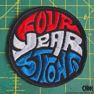 May include: A circular embroidered patch with the words "FOUR YEAR STRONG" in red, white, and blue. The patch has a black border and is on a green surface with grid lines.
