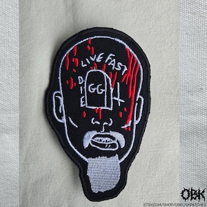 Puede incluir: Parche bordado negro con una cara estilizada, las palabras "LIVE FAST" y "DIE" sobre una lápida con "GG". Acentos rojos que se asemejan a la sangre. El parche tiene un contorno blanco y una barba blanca.