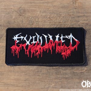 May include: A rectangular black embroidered patch featuring the word EVILIVED in a stylized white font. The lower portion of the lettering is accented with red embroidery that appears to drip downward, creating a distinct visual effect on the fabric.