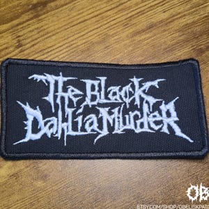 Pode incluir: Um emblema retangular bordado em preto com o texto branco The Black Dahlia Murder numa fonte estilizada e irregular. O emblema tem uma borda preta espessa costurada e foi concebido para ser aplicado em superfícies de tecido.