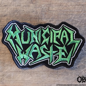 May include: A black embroidered patch featuring the text Municipal Waste in a jagged, stylized green font with a white border. The lettering is sharp and angular, set against a solid black fabric background with a matching black stitched edge.
