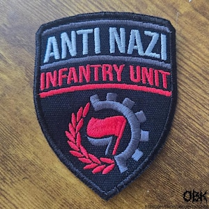 Puede incluir: Un parche bordado en forma de escudo con fondo negro. Presenta el texto ANTI NAZI en gris claro e INFANTRY UNIT en rojo. Debajo del texto hay un símbolo de bandera roja rodeado por un engranaje gris y una corona de laurel roja.