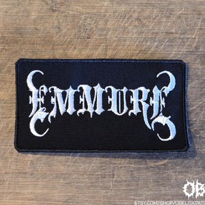May include: A rectangular black embroidered patch featuring the white text EMMURE in a stylized, jagged gothic font. The patch has a black stitched border and is designed for application to fabric items like clothing or bags.