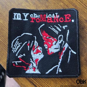 May include: Black square patch with the text "MY CHEMICAL ROMANCE" in white and red. The patch features a stylized graphic of two figures in white and red on a black background. The patch is on a wooden surface.