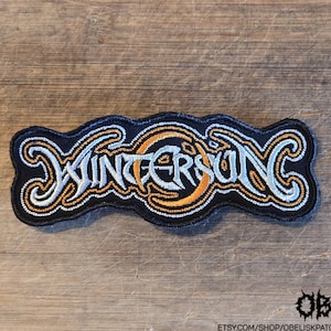 Puede incluir: Un parche bordado que muestra la palabra Wintersun en una fuente estilizada de color blanco y azul claro, con una forma de media luna naranja integrada en el diseño, todo sobre un fondo negro sólido con un borde negro grueso.