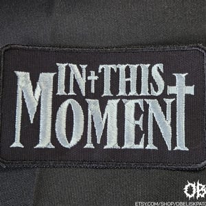 May include: Black rectangular patch with the words "IN+THIS MOMENT" in silver lettering. The patch has a cross design incorporated into the text. The patch is likely for clothing or accessories.