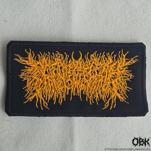 May include: Black rectangular patch with yellow embroidered abstract design. The patch has a textured appearance and features the text "OBK" in the bottom right corner, along with a website address.