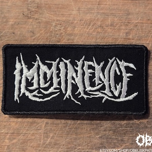 May include: A rectangular black embroidered patch featuring the word Imminence in a white, jagged, stylized font. The embroidery has a dense texture with a black border surrounding the white lettering, creating a high-contrast design.
