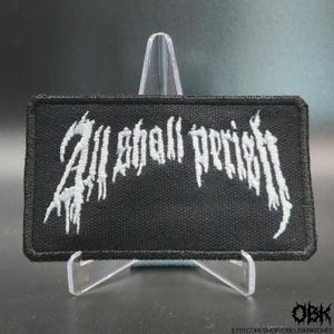 Puede incluir: Un parche rectangular bordado en negro con el texto blanco All Shall Perish en una fuente estilizada, dentada y desgastada. El parche tiene un borde grueso cosido en negro y se muestra sobre un soporte de plástico transparente.