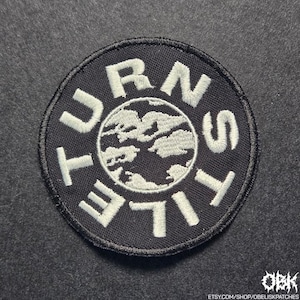 Pode incluir: Um patch redondo preto com texto bordado branco e um desenho central. O texto diz "TURNSTILE" em um padrão circular em torno de um gráfico branco de um globo estilizado. O patch é provavelmente para roupas ou acessórios.