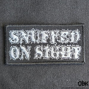 May include: A rectangular black patch with white embroidered text that reads "SNIFFED ON SHIT". The patch has a white border and is set against a dark gray fabric background. The text is in a stylized, gothic font.
