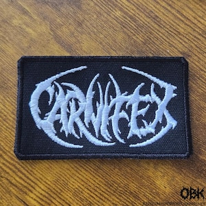 May include: A rectangular black embroidered patch featuring the white text Carnifex in a stylized, jagged, death metal-inspired font. The patch has a thick black stitched border and a textured fabric base, with the text centered on the surface.