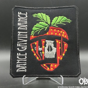 Könnte beinhalten: Schwarzer, gestickter Aufnäher mit dem Text "DANCE GAVIN DANCE" vertikal auf der linken Seite. Der Aufnäher hat ein Erdbeer-Design mit einem Totenkopf in der Mitte. Die Erdbeere ist rot mit gelben Punkten und grünen Blättern.