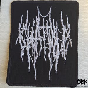 May include: Black square patch with white embroidered text in a gothic font. The patch has a black border and a white background. The text appears to be a band logo or design. The patch is likely for a jacket, bag, or other apparel.