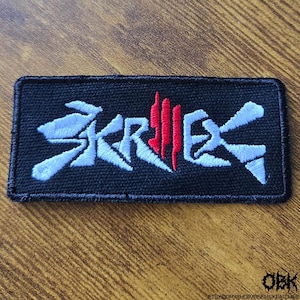Può includere: Toppa rettangolare nera con la scritta "SKRILLEX" ricamata in bianco e rosso. La toppa ha un bordo nero ed è probabilmente un accessorio per abbigliamento o borse. Il testo è stilizzato con un carattere unico.