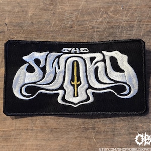 May include: Black rectangular patch with white embroidered text "THE SWORD" and a gold sword design. The patch has a black border and is likely for clothing or accessories.