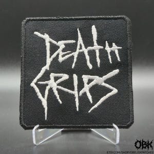 Puede incluir: Un parche cuadrado bordado en negro que presenta el texto blanco Death Grips en una fuente dentada y estilizada. El parche tiene un borde grueso cosido en negro y se muestra sobre un soporte de plástico transparente, resaltando el detalle.