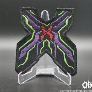 Könnte beinhalten: Schwarzer, gestickter Aufnäher in X-Form mit lila, grünen und roten Akzenten. Der Aufnäher hat ein detailliertes Design mit strukturiertem Aussehen. Der Aufnäher wird auf einem transparenten Ständer präsentiert.