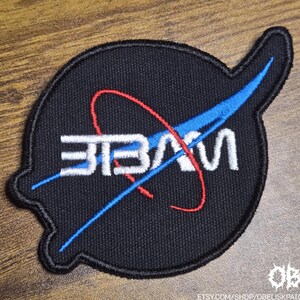 Può includere: Toppa nera con design "NASA" bianco, un arco rosso e un elemento blu. La toppa ricamata ha una forma distintiva, adatta per abbigliamento o accessori.